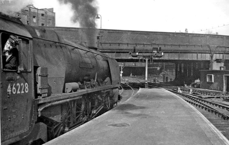 BloodandCustard Euston Station 1962
Platform 13/14
Departure side: outward at the end of Platform 13, Stanier 'Coronation' 8P no. 46228 'Duchess of Rutland' is about to leave on the 10.25 to Carlisle and Windermere.
Ahead is the Ampthill Square Bridge No. 2. Until 1952 there would also have been the Bridge No. 1, making the whole top end of the Station very restricted and claustrophobic, dominated by the great No. 2 Signalbox; No. 2 Bridge was also removed when the New Euston was built.
Ben Brooksbank (CC-by-SA/2.0)