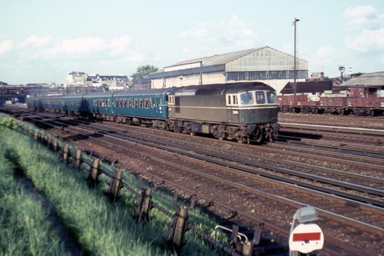 Against the backdrop of Wimbledon’s S&T depot (the former Flying Boat shed from Newhaven) and sporting the pre-10th July 1967 Basingstoke ‘39’ headcode D6544 loco-hauls TC stock forming the 18.09 Waterloo – Basingstoke service on 26th May 1967. Although the leading unit is running as a 3TC, it is numbered as a 4TC.
© A Noakes (R Carroll Collection)
