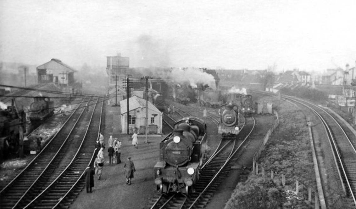 Serving Norwood Marshalling Yard, Norwood Depot was situated on the east side of the main line north of Norwood Junction station, with the single-track Down Crystal Palace Spur from Clapham Junction (via Crystal Palace and Bromley Junction) curving round on the right. 
In November 1954 the depot (BR code 75C) had an allocation of 33 steam locomotives (17 0-6-0, 4 2-6-4T, 12 0-6-2T) and 14 Diesels (1 main-line, 13 shunters), most of which remained in early 1960. The 'civilians' wandering around are on an RCTS visit on Saturday, 12th March 1960.
© Ben Brooksbank (Geograph/CC-by-SA)
